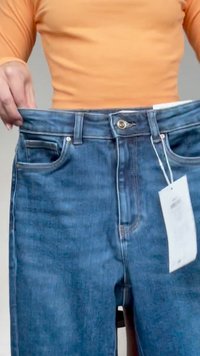 Person i oransje topp holder blå jeans ut ved midjen, og viser løs passform med hvitt prislapp festet i knappområdet.
