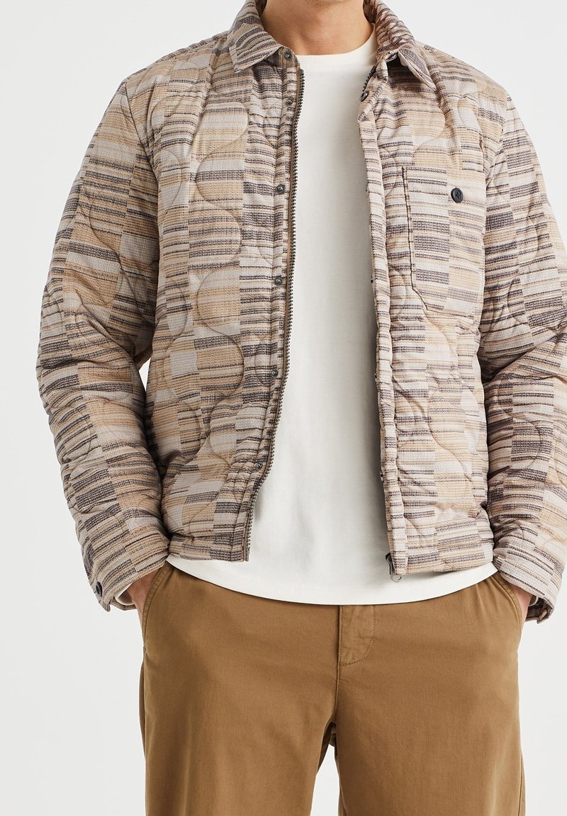 Mann trägt eine beige-braun gemusterte gesteppte Jacke über einem weißen Hemd, die Hände in den Taschen der braunen Hose, Bildausschnitt an Hals und Oberschenkeln.