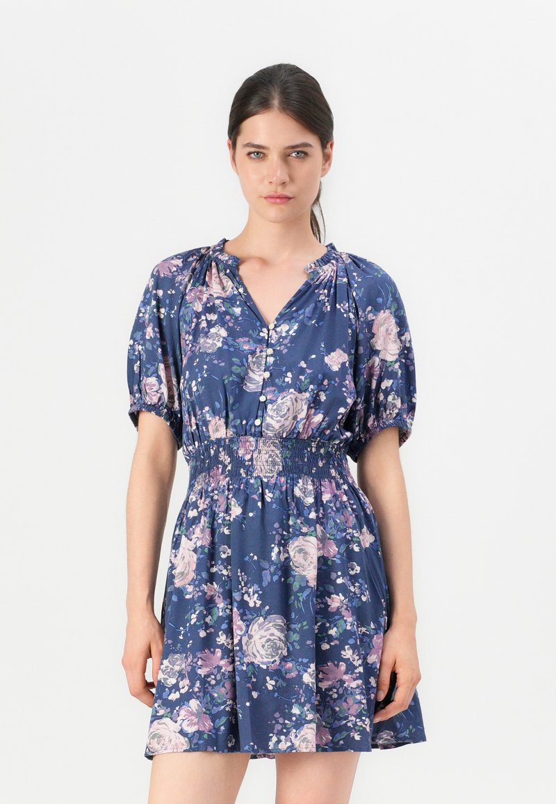 Robe fleurie en tissu bleu marine, avec des manches courtes bouffantes, un col en V et une taille élastique. Motifs floraux dans des tons rose, blanc et vert.