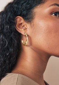 Gold hoop earring with a twisted design shown on a textured, dark hair background. The earring features a shiny finish and open shape.
