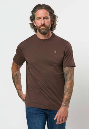 Man met krullend haar en baard, draagt een bruin T-shirt en blauwe spijkerbroek, toont getatoeëerde armen tegen een effen lichte achtergrond.