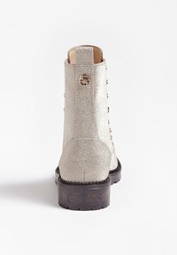 Bottine argentée brillante avec des lacets sur le côté, bouton logo en métal rond, et semelle noire embossée avec "GUESS" vue de l'arrière.