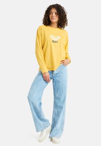 Żółta bluzka z długim rękawem z białym logo, zestawiona z jasnoniebieskimi spodniami typu wide-leg i białymi sneakersami. Casulowy, luźny krój.