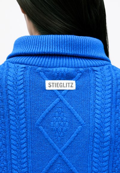 Suéter azul de punto con un patrón texturizado que presenta franjas y motivos de diamante, complementado por un cuello doblado y una etiqueta que dice "STIEGLITZ."