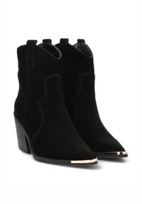 Botines de ante negros hasta el tobillo con punta afilada, tacón apilado y acentos metálicos en la punta. Incluyen lengüetas para facilitar el uso.