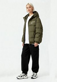 Doudoune vert olive avec fermeture éclair à l'avant, capuche, et motif blanc de cœur et flèche ; associée à un pantalon cargo noir et des baskets blanches.