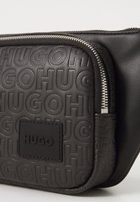 Borsetta nera in pelle con motivo in rilievo con testo "HUGO" ripetuto, una cerniera frontale e un'etichetta nera cucita con il logo "HUGO" al centro.