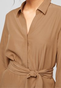 Vestito avvolgente tan con maniche lunghe, colletto e cintura legata in vita. Tessuto liscio con una leggera lucentezza e dettagli di cucitura puliti.