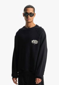 Černá mikina s volným střihem, s raglánovými rukávy a stříbrným vyšívaným logem na prsou. Hladký povrch, minimalistický design.