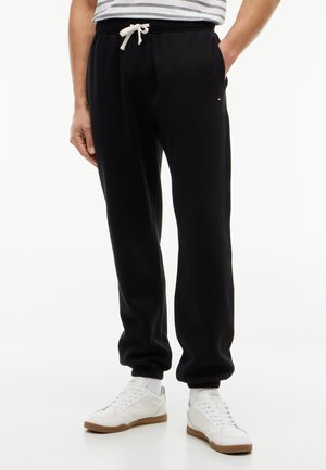 Homme portant un pantalon de jogging noir avec un cordon blanc et des baskets blanches, debout sur fond blanc.