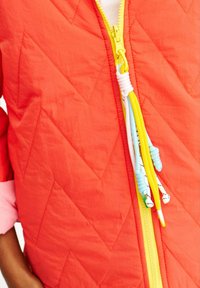 Světlá oranžová prošívaná bunda se žlutým zipem a barevnými tkaničkami v modrých, žlutých a bílých vzorech.