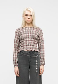 Νεαρό άτομο με μακριά ξανθά μαλλιά, που φοράει μπλούζα με καρό μοτίβο και σφηκωτή ύφανση, σκουρόχρωμο παντελόνι με φαρδιά μπατζάκια και διακοσμητική ασημένια αλυσίδα με αστεράκια ως ζώνη.