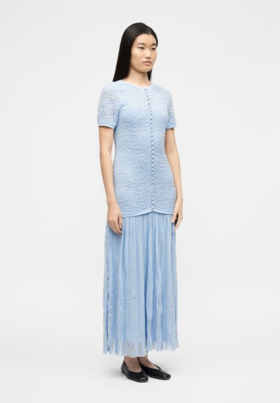 Γυναίκα με ίσια μαύρα μαλλιά, φορώντας ένα ανοιχτό μπλε δαντελωτό φόρεμα με κουμπιά στο μπροστινό μέρος και μαύρες επίπεδες παπούτσια, που στέκεται μπροστά σε μια απλή φόντο.