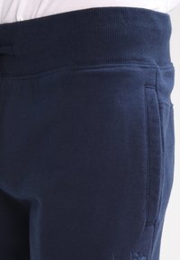 Pantalones deportivos azul marino con cinturilla acanalada, bolsillos laterales y detalle de logotipo bordado. Material de algodón suave con una textura lisa.
