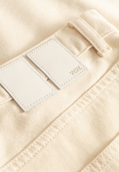 Zbliżenie na beżowe spodnie materiałowe z podwójnie białymi naszywkami, z których jedna ma wytłoczone litery "WW." obok szlufki na pasek i szwu.