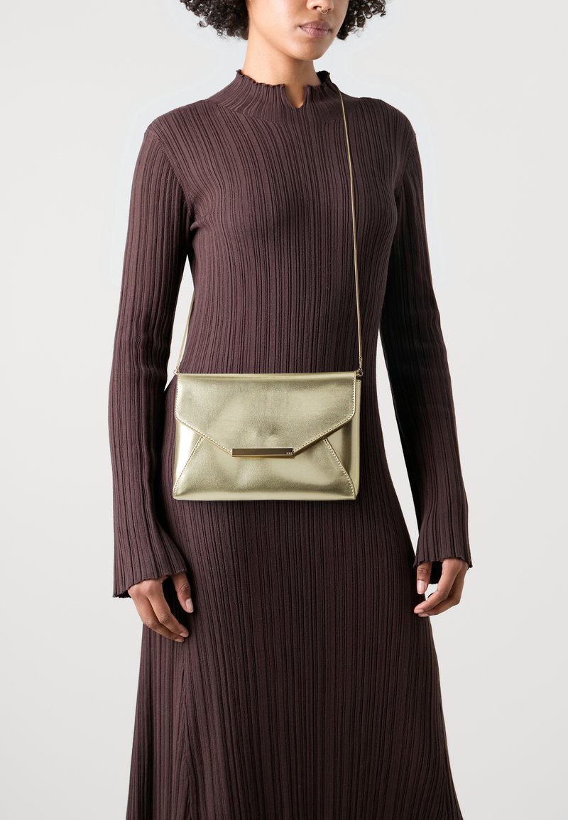 Sac à bandoulière doré métallisé avec un design en forme d'enveloppe, doté d'une finition texturée et d'un accent matériel minimaliste et plat. Porté sur une robe brune à côtes.