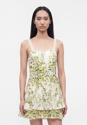 Femme aux longs cheveux foncés porte une robe blanche sans manches avec une broderie florale jaune et verte, debout devant un fond clair uni.