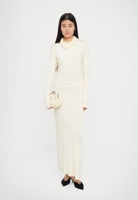 Abito a maniche lunghe di colore crema con una silhouette aderente, tessuto strutturato, scollo a barchetta e una borsa a pochette coordinata. Scarpe nere completano il look.
