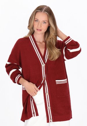 Kvinde iført en bordeaux cardigan med hvide striber og knapper, poserer med den ene hånd i lommen og den anden, der rører ved hendes hår.