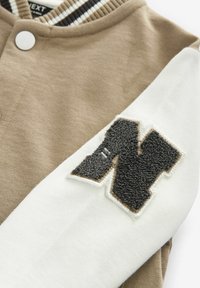 Manche beige et blanche d’une veste universitaire avec un écusson lettre « N » noire texturée cousu sur le tissu blanc.