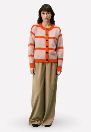 Femme portant un cardigan boutonné rayé rose, gris et orange, et un pantalon large beige, debout les bras détendus le long du corps.