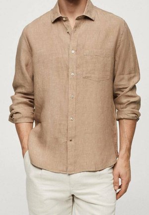 Homme portant une chemise beige à manches longues boutonnée avec manches retroussées et un pantalon blanc, mains partiellement dans les poches, fond neutre.