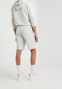 Personne portant un sweat à capuche gris clair et un short assorti, des chaussettes blanches et des baskets blanches, se tenant de dos contre un fond blanc uni.