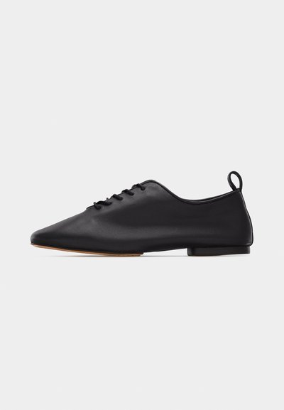 Chaussure plate en cuir noir à lacets, avec un bout pointu et un petit talon, dotée d'une languette à l'arrière, vue de profil sur un fond blanc.