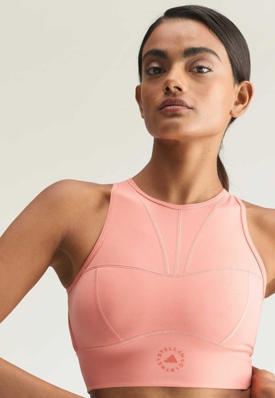Woman with dark hair in a low ponytail wearing a fitted pink athletic crop top with a round neckline against a plain light background.