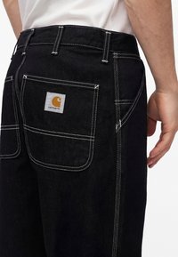 Osoba nosiaca čierne nohavice Carhartt s výrazným bielym prešívaním a viditeľnou nášivkou s logom na zadnom vrecku na bielom pozadí.