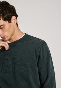 Sudadera verde oscura con un logotipo texturizado, cuello redondo y puños acanalados. Tejido suave con un corte relajado.