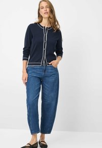 Marineblauer Cardigan mit weißer Paspelierung, Knopfverschluss und langen Ärmeln, kombiniert mit locker sitzenden blauen Jeans und schwarzen Ballerinas. Durchgehend glatte Textur.