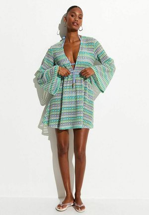 Femme portant une robe courte à manches longues à motifs avec des manches évasées, nouée à la taille, debout devant un fond blanc.