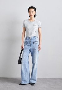 Tricou gri cu mânecă scurtă și logo-ul EA7, asortat cu blugi largi de culoare albastru deschis, cu buzunare frontale și fermoar central. Ține o geantă neagră.