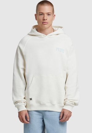 White hoodie with a front pocket, ribbed cuffs, and a small light blue logo. The fabric appears soft and textured, paired with light blue jeans.
