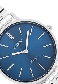 Montre en acier inoxydable argenté avec un cadran rond bleu, des index d'heure fins argentés et des aiguilles blanches. Comprend le texte "OOZOO" et "Vintage".