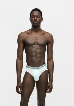 Modèle masculin avec une silhouette tonique portant un slip blanc Calvin Klein, debout devant un fond uni gris clair, regardant devant.
