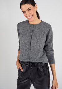 Cardigan en laine grise avec des poignets côtelés, doté d'un col rond et d'une fermeture à boutons. Associé à un pantalon à cordon en cuir noir.