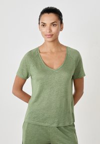 Woman with dark hair tied back wearing a light green short-sleeve V-neck top and matching pants, standing with hands behind back.