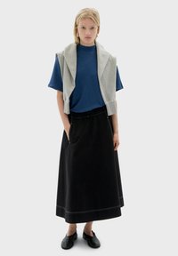 Haut bleu à manches courtes avec un pull gris posé sur les épaules, associé à une longue jupe noire et des chaussures noires. Couture notable sur la jupe.