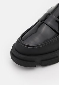 Mocassin en cuir noir brillant avec une semelle en caoutchouc épaisse et texturée, bout arrondi et une bride sur le dessus, photographié sur fond blanc.