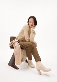 Beige Langarmoberteil, olivgrüne Hose, kombiniert mit einem hellbeigen Trenchcoat. Strukturierte cremefarbene Stiefel mit blockierendem Absatz. Brauner Stuhl.