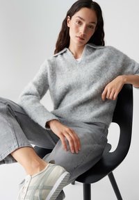 Grijze donzige pullover met een kraag, gecombineerd met lichtgrijze denim broek en off-white sneakers met groene accenten, zittend op een zwarte stoel.