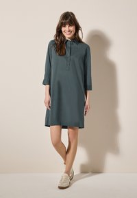 Abito camicia verde scuro con colletto e patta con bottoni. Presenta maniche a tre quarti e una vestibilità ampia, abbinato a scarpe da ginnastica beige.