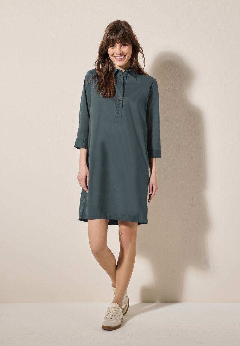 Abito camicia verde scuro con colletto e patta con bottoni. Presenta maniche a tre quarti e una vestibilità ampia, abbinato a scarpe da ginnastica beige.