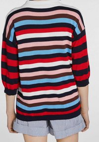 Multikleurige gestreepte trui in rood, blauw, pink en wit, met een ronde halslijn en losse, opbollende mouwen. Draagt over gestreepte shorts.