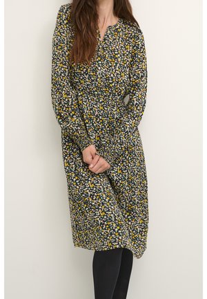 Femme portant une robe midi à manches longues avec des fleurs noires, jaunes et blanches, taille froncée, debout devant un fond clair uni.