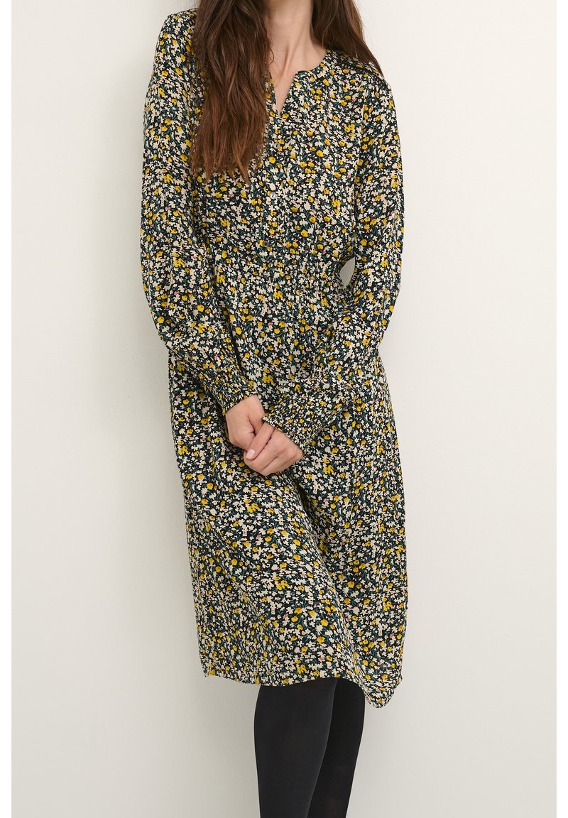 Femme portant une robe midi à manches longues avec des fleurs noires, jaunes et blanches, taille froncée, debout devant un fond clair uni.