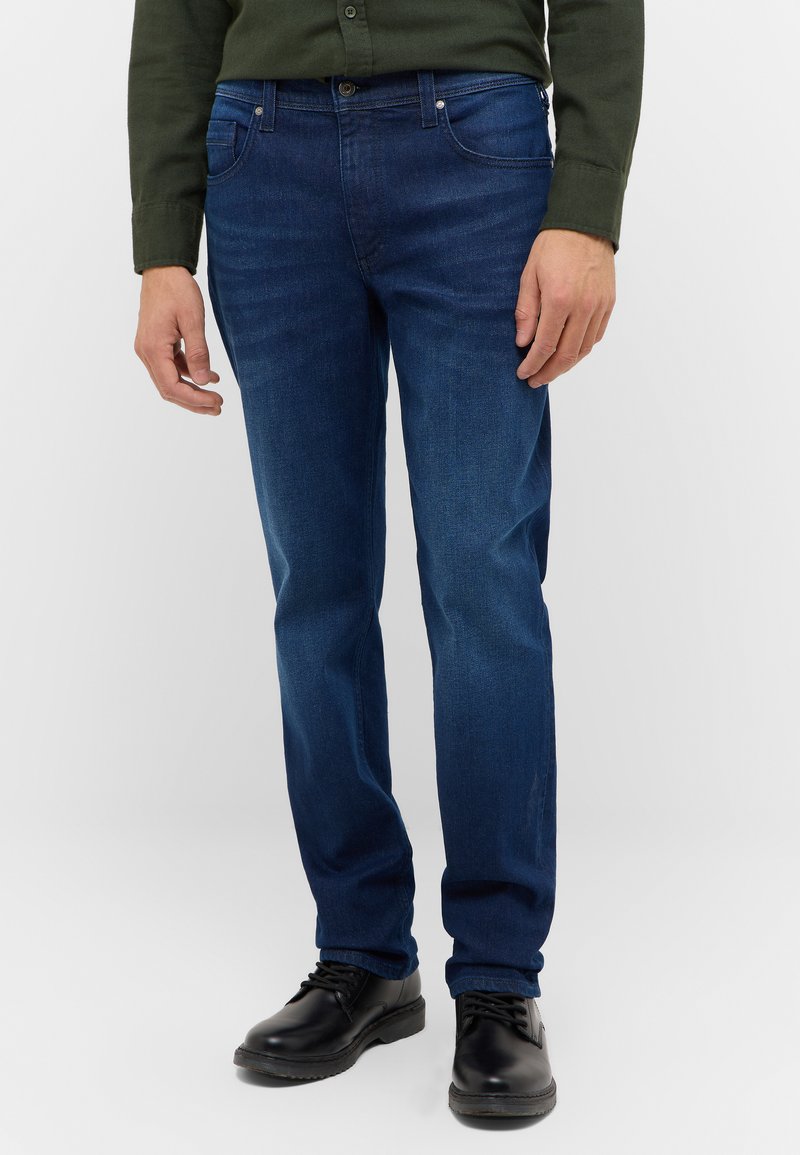 Donkerblauwe jeans met een rechte snit, voorzien van voorzakken en een knoopsluiting. Gekweld met zwarte schoenen en een groene shirt.
