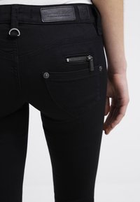 Jeans de mezclilla negra con un diseño ajustado, que cuentan con un bolsillo con cremallera y detalles de costura en la parte posterior. Etiqueta de cuero en la cinturilla.